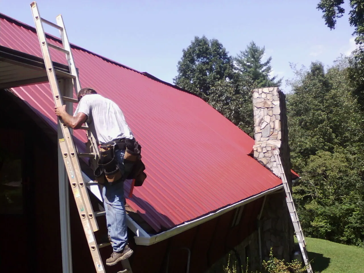 Roofer on a ladder beside a red metal roof and stone chimney — Soffit & Fascia Repair by Roofing Brothers in Hartford